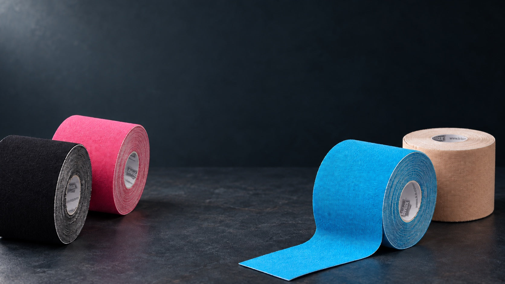 K-Tape-Rollen in Blau, Beige, Pink und Schwarz auf dunklem Untergrund.